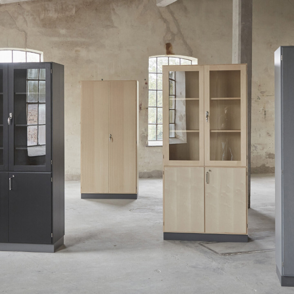 Fyra fristående skåp är arrangerade i ett stort tomt rum med betonggolv och exponerade tegelväggar. Fönster i bakgrunden tillåter naturligt ljus att lysa upp utrymmet. Skåpen varierar i färg, med svarta och ljusa träfinisher.
