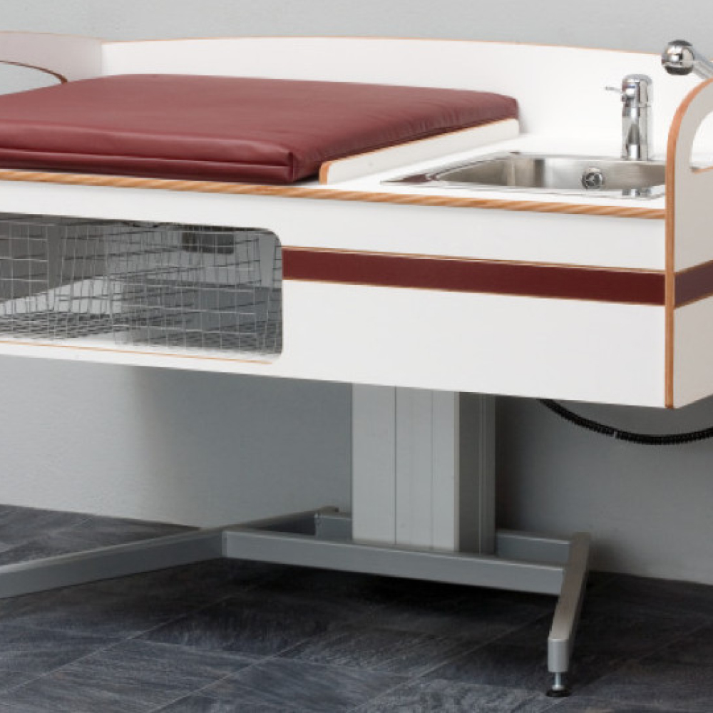 Ett vitt veterinärundersökningsbord med en rödbrun kudde och en inbyggd diskbänk i rostfritt stål. Bordet har ett trådförvaringsfack undertill och står på ett grått klinkergolv.