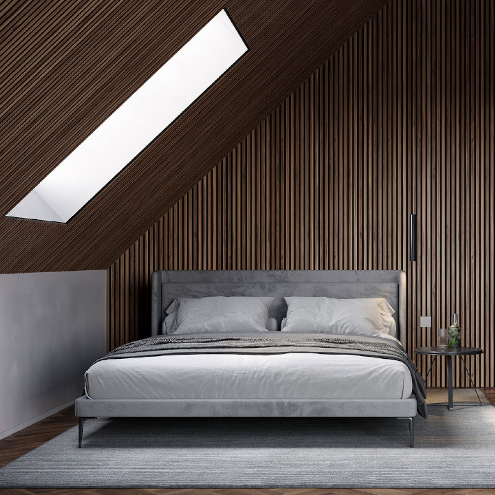 Ett modernt sovrum med ett takfönster som visar en lutande träpanelvägg. En grå stoppad säng med vita och grå sängkläder är centrerad på en grå matta. Ett litet sidobord med en svart lampa finns till höger.