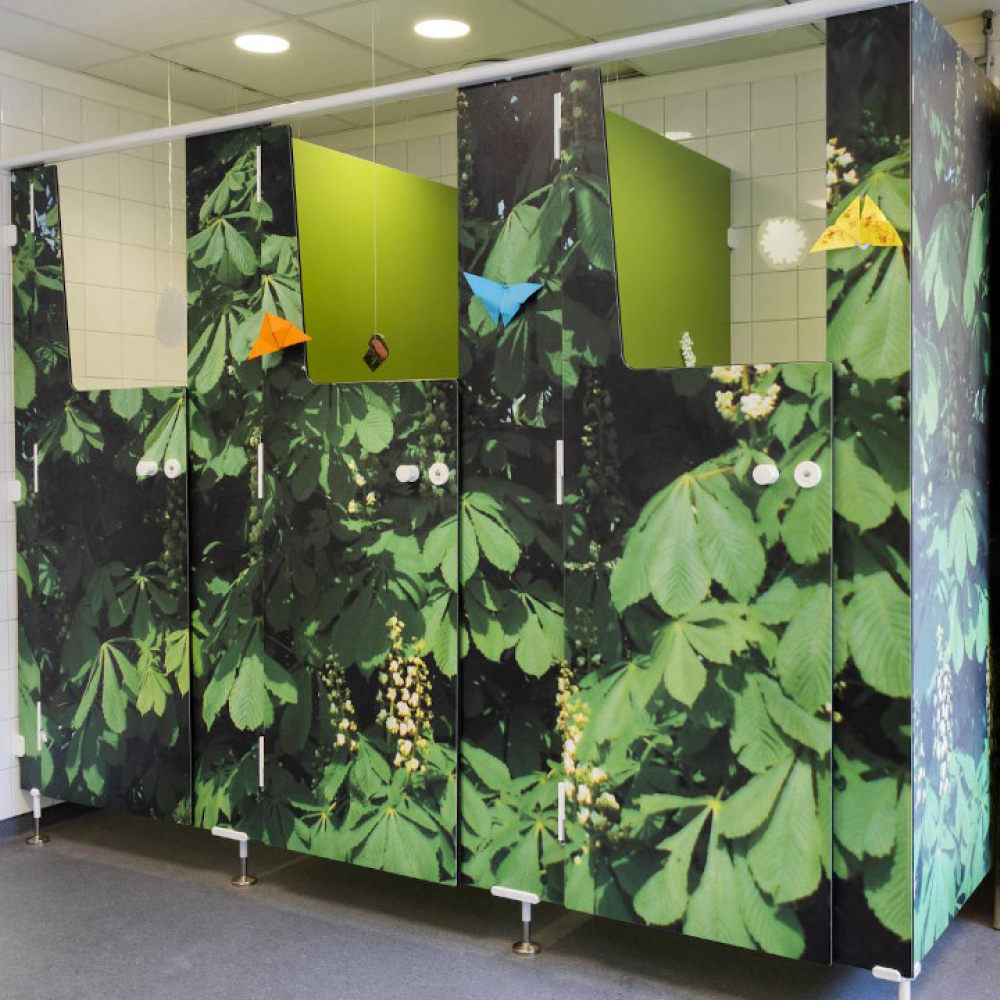 Badrumsstånden har en livlig design med skogstema med frodigt grönt bladverk. Dörrarna är prydda med pappersorigami i olika färger. Golvet är grått och taklampor lyser upp utrymmet.