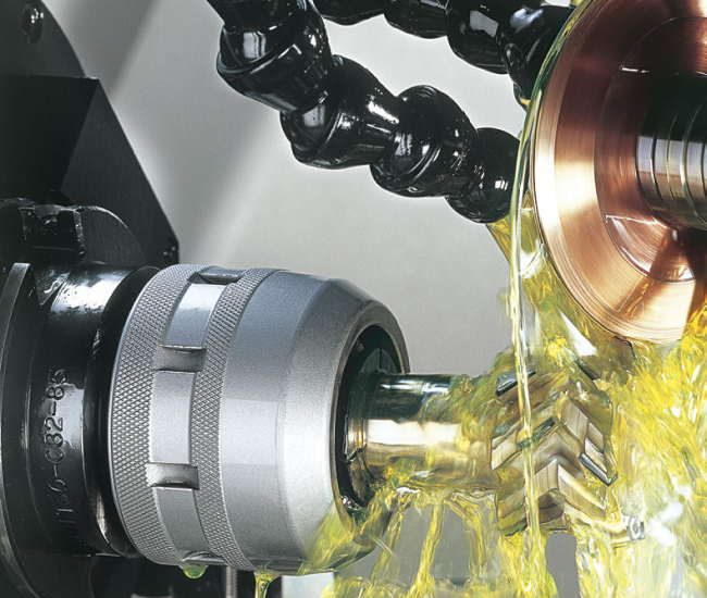 Närbild av en metallbearbetningsmaskin med kylvätska som strömmar över ett roterande kugghjul som bearbetas. Installationen inkluderar en cylindrisk komponent och en svart slang som leder vätskan. Scenen visar industriell precisionsbearbetning på gång.