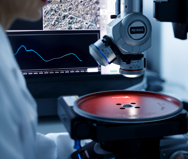 En laboratoriemiljö med ett mikroskop som undersöker ett prov på en röd skiva. En monitor visar ett grafiskt linjediagram och en gråskalebild. En person i vit rock är delvis synlig, med fokus på utrustningen.