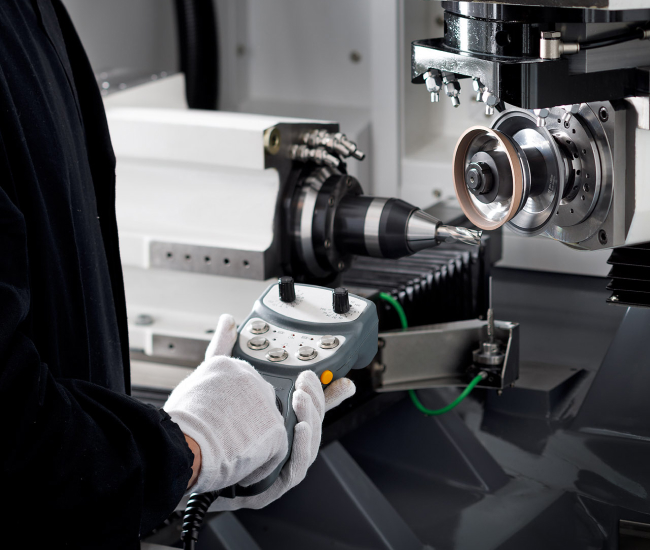 En person i vita handskar styr en kontrollpanel för en precisionsmaskin. Maskinen, med metalldelar och en slipskiva, används sannolikt för att skära eller forma material.