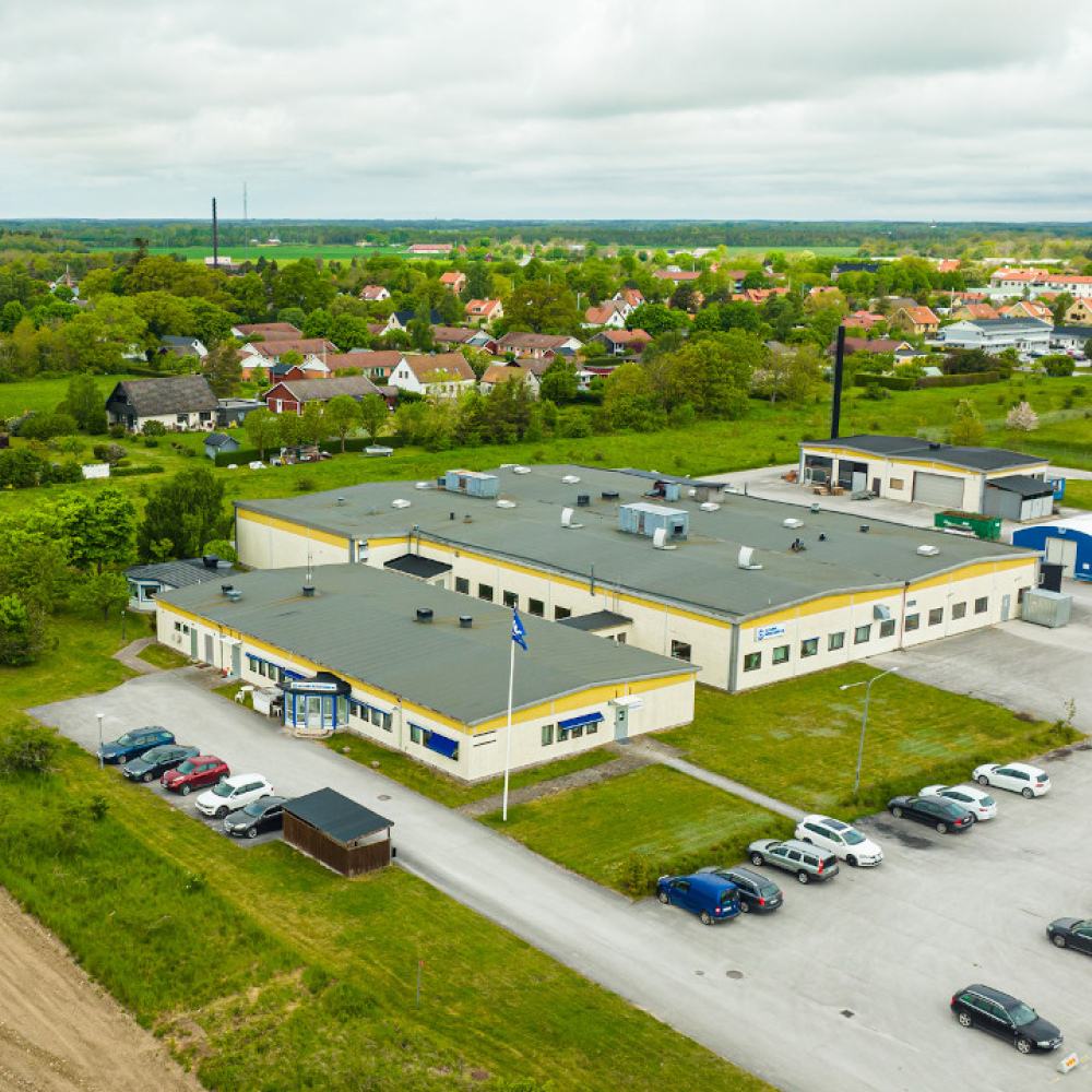 Flygfoto över en stor industribyggnad med gula accenter, omgiven av grönska och några parkerade bilar. I bakgrunden syns ett bostadsområde med utspridda hus och avlägsna träd under en mulen himmel.